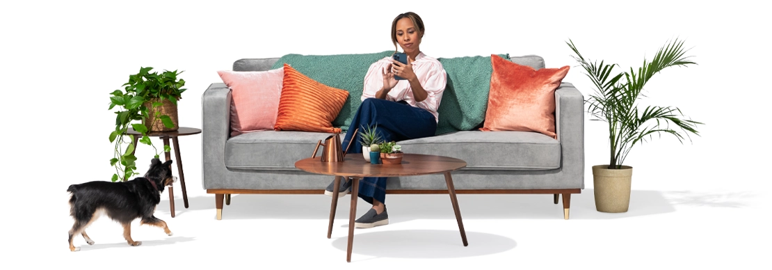 As her dog approaches, a young woman on a grey couch between two potted plants and colored pillows uses her smart phone to check out how much she could save by switching to State Farm.